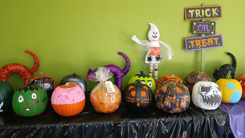 A long view of the pumpkin display table against a green wall, featuring painted pumpkins designed as a green monster face, a pink pastry with sprinkles, a cat face, and a patterned design, flanked by sparkly tinsel cat silhouettes and a ghost decoration.