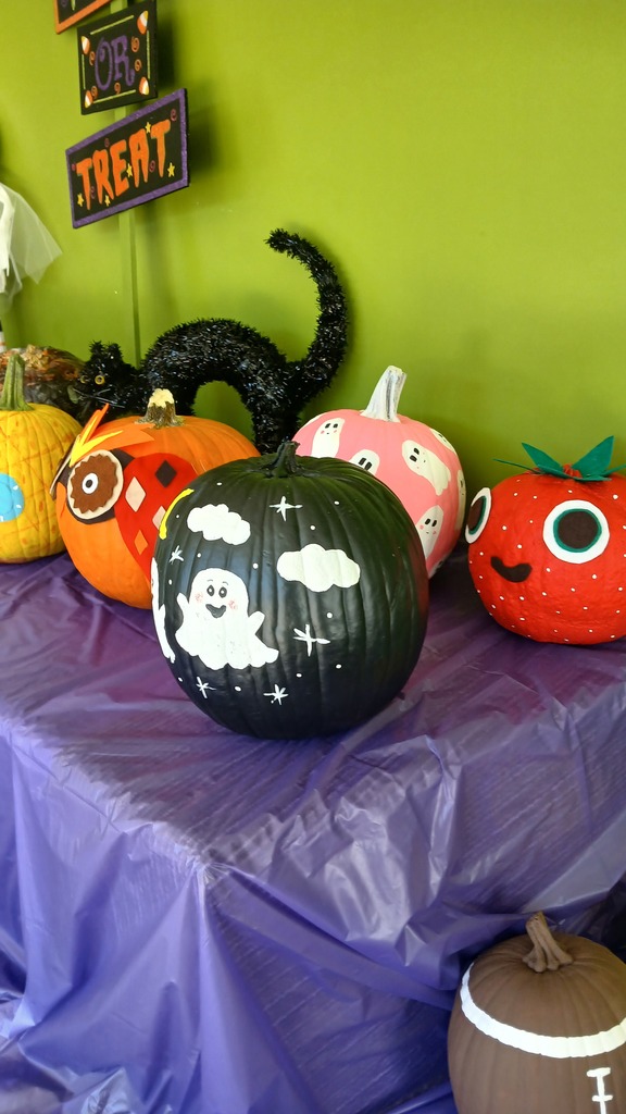 A close-up view of painted pumpkins on a purple table covering, featuring a black pumpkin decorated with a white ghost and stars, a pink pumpkin with tiny white ghosts, a strawberry-themed pumpkin, and an owl-decorated pumpkin.