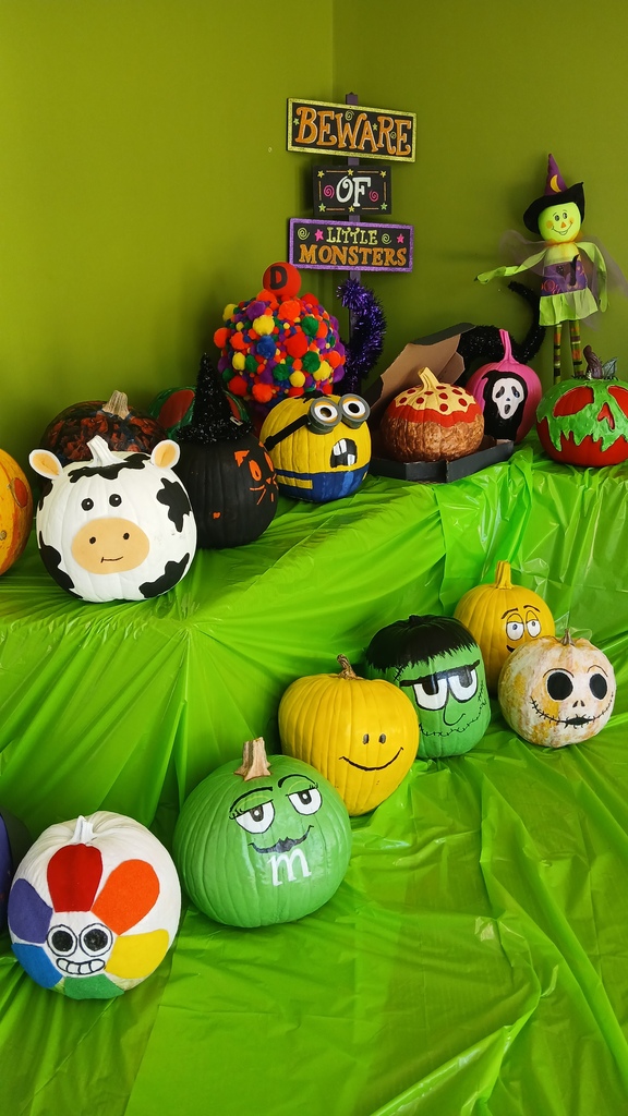 A colorful display of creative, parent-painted pumpkins set against a bright green wall on a table covered with a green plastic tablecloth, featuring character designs like a cow, a Minion, a green monster, and a colorful pom-pom-covered creation.