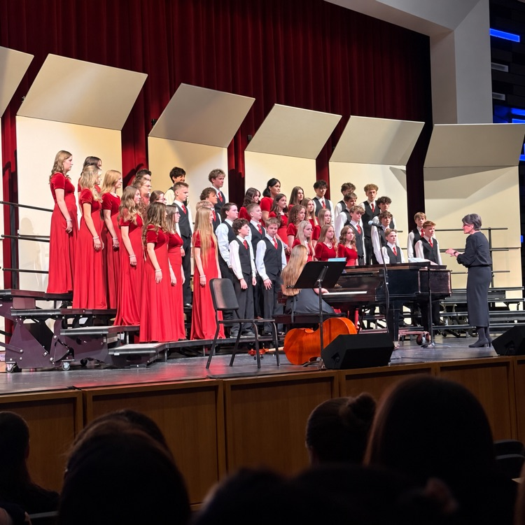 Choir students in red dresses and black vests perform under the direction of their conductor.