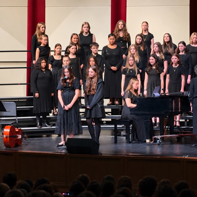 Two choir students stand at a microphone with their choir and pianist behind them.
