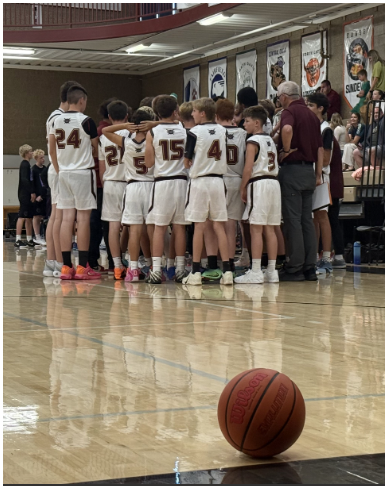 Alt Text: A boys basketball team huddles with coaches on a gym floor during a game or timeout, while a basketball rests in the foreground. Banners hang on the wall behind the group, and the polished court reflects the players and gym lights.