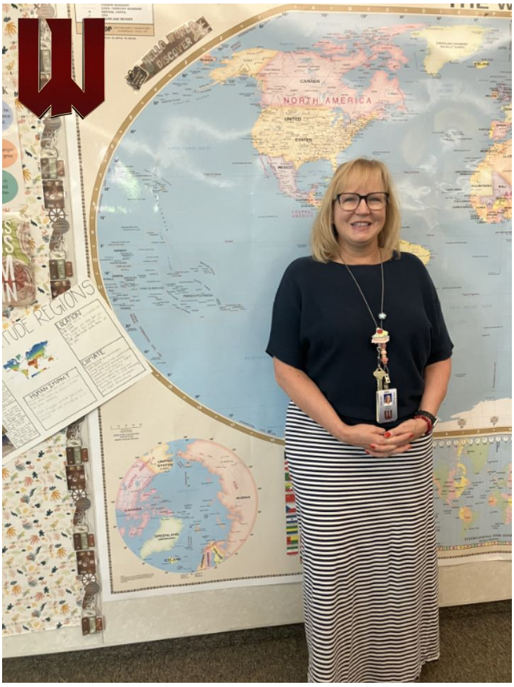 A woman wearing a black shirt and a black and white striped skirt. She is standing in front of a map in her classroom.