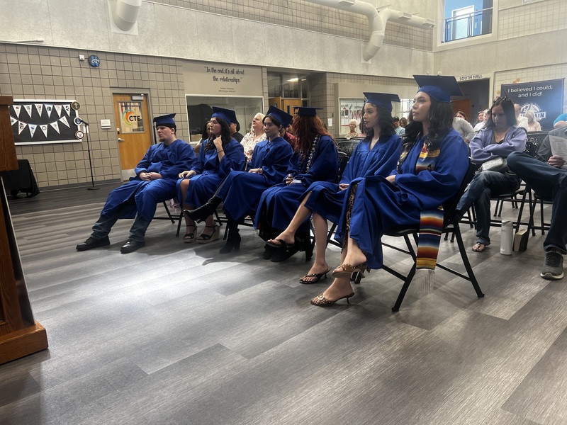 6 students on the front row dressed in graduation gowns watching a speaker in front of them