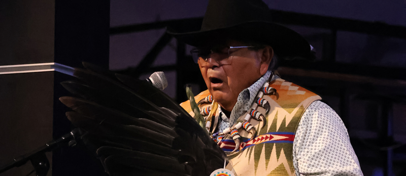 A man dressed in traditional Native American clothing sings while holding a black feather.