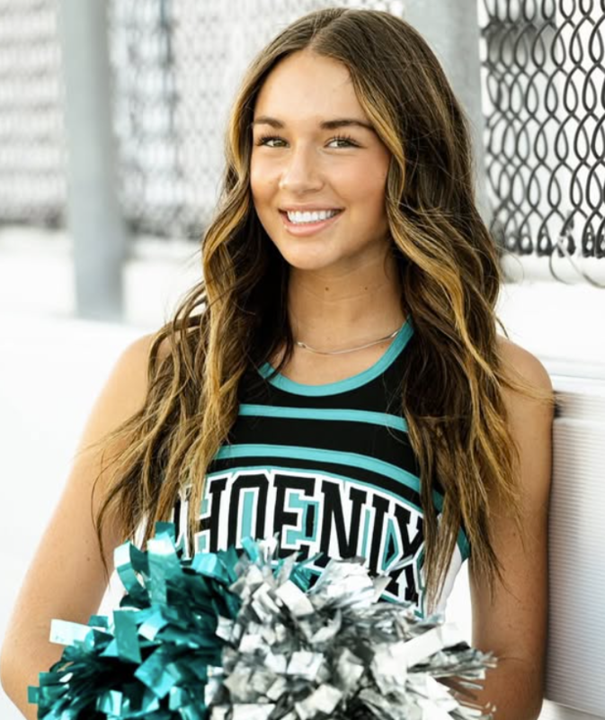 A cheerleader with teal and silver pom poms in a teal, white, and black uniform smiling at the camera with bleachers behind them