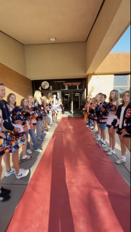 I line of students going down both sides of red butcher paper set on the ground to represent a red carpet.  There cheerleaders in their white, orange and blue uniforms along with school officers in their blue, orange and white sweaters. 