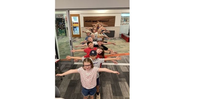 A class of students face the camera in a line to the far wall, arms outstretched under a “What’s Your Wingspan?” sign marked with wingspans, including a bald eagle’s.