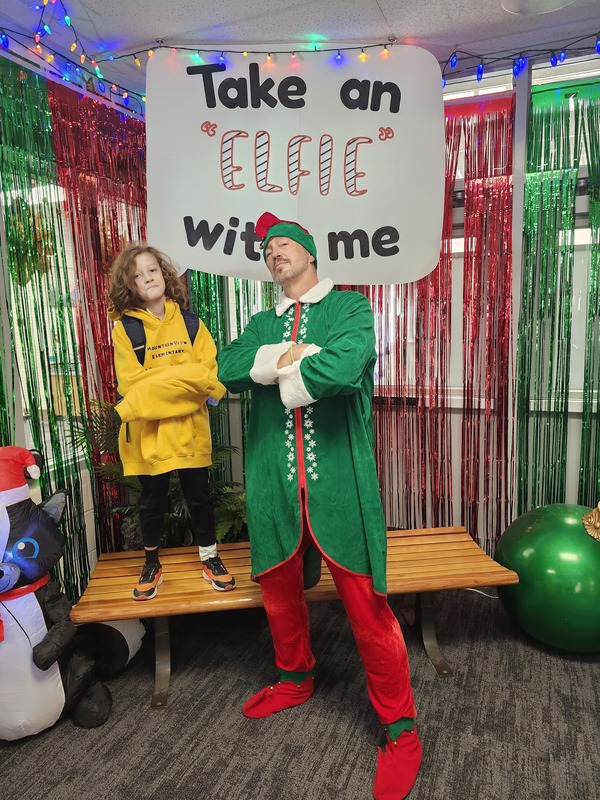 Children posing with Pinkie the Elf in front of decorations and a sign that says Take an "Elfie" with me.
