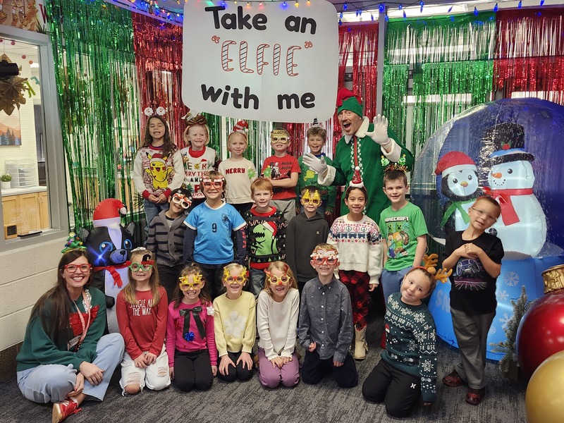 Children posing with Pinkie the Elf in front of decorations and a sign that says Take an "Elfie" with me.