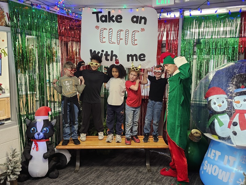 Children posing with Pinkie the Elf in front of decorations and a sign that says Take an "Elfie" with me.
