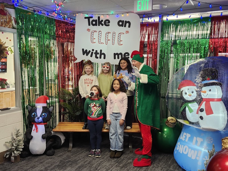 Children posing with Pinkie the Elf in front of decorations and a sign that says Take an "Elfie" with me.