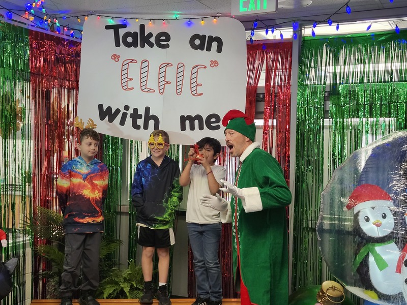 Children posing with Pinkie the Elf in front of decorations and a sign that says Take an "Elfie" with me.