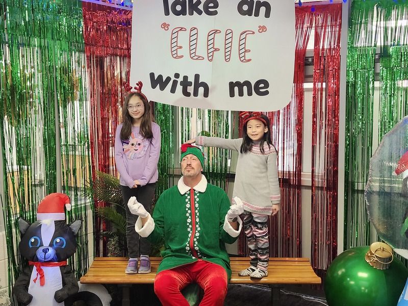 Children posing with Pinkie the Elf in front of decorations and a sign that says Take an "Elfie" with me.
