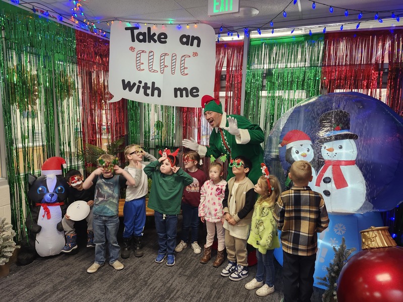 Children posing with Pinkie the Elf in front of decorations and a sign that says Take an "Elfie" with me.
