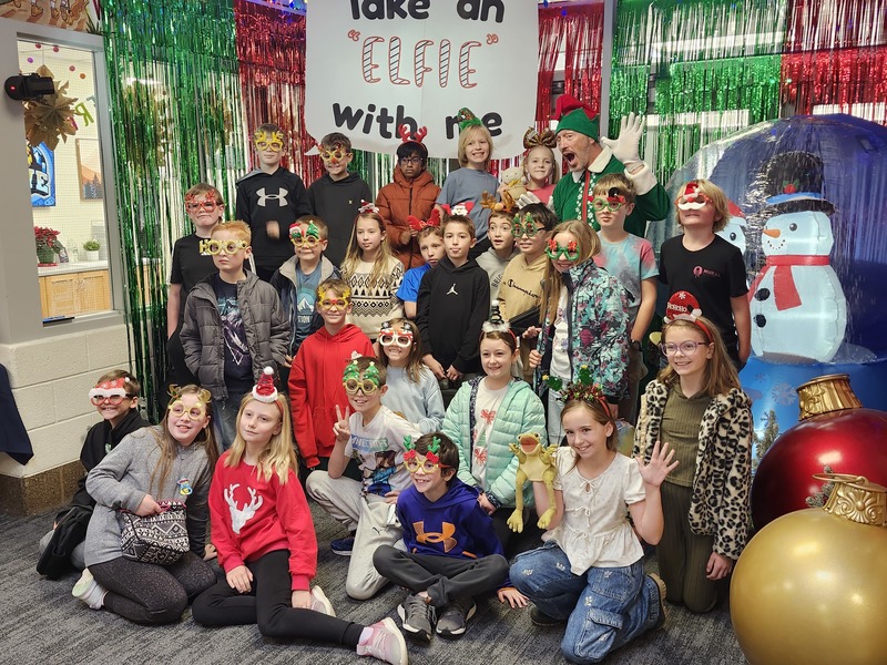 Children posing with Pinkie the Elf in front of decorations and a sign that says Take an "Elfie" with me.