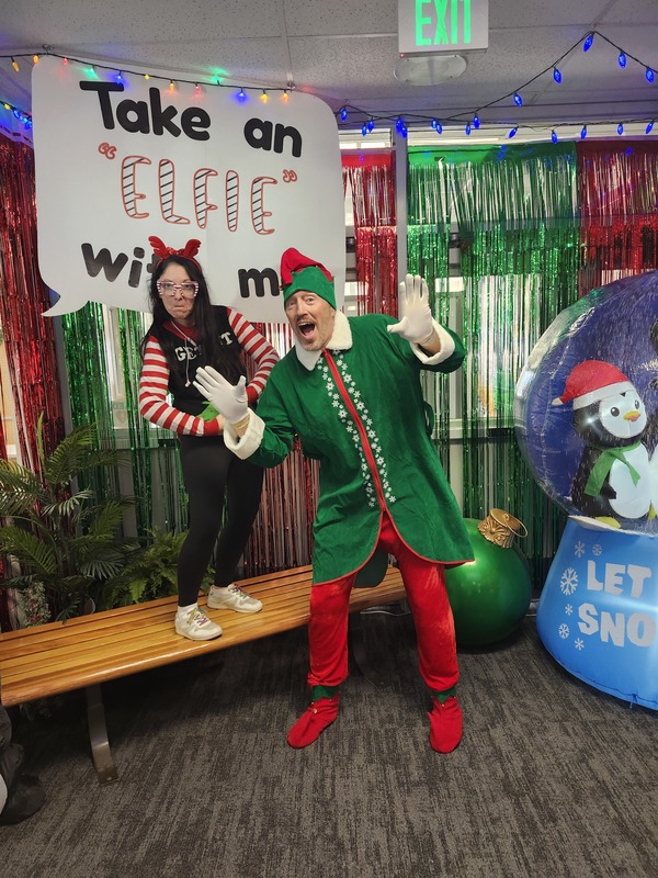 A parent posing with Pinky in front of a sign that says Take an "Elfie" with me.