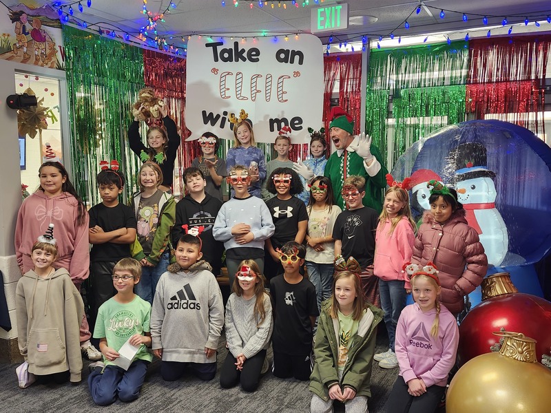 Children posing with Pinkie the Elf in front of decorations and a sign that says Take an "Elfie" with me.