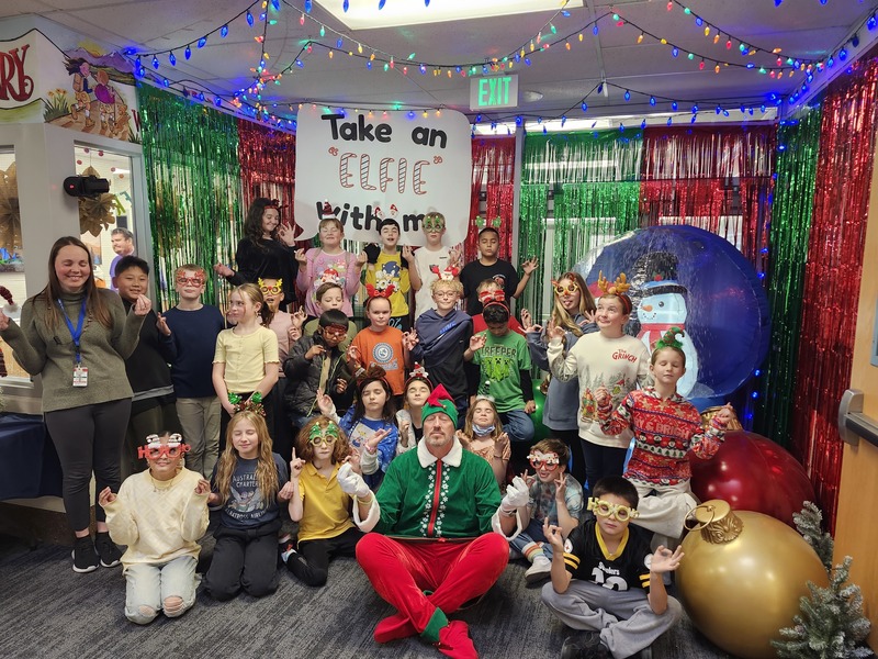 Children posing with Pinkie the Elf in front of decorations and a sign that says Take an "Elfie" with me.