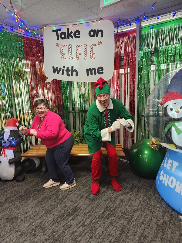 Teacher posing with Pinky the Elf in front of a sign that says Take an "Elfie" with me.