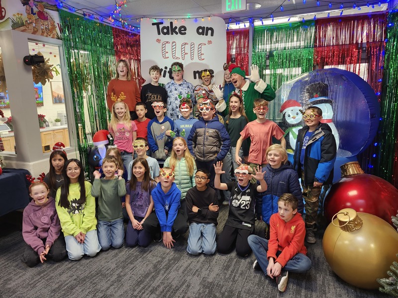 Children posing with Pinkie the Elf in front of decorations and a sign that says Take an "Elfie" with me.