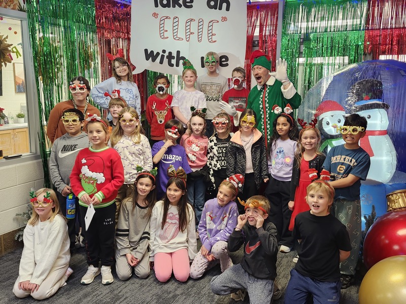 Children posing with Pinkie the Elf in front of decorations and a sign that says Take an "Elfie" with me.