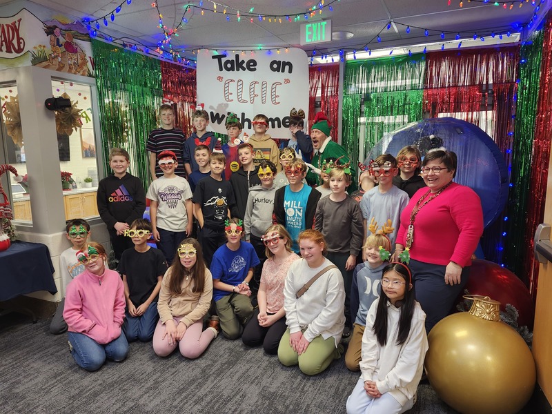 Children posing with Pinkie the Elf in front of decorations and a sign that says Take an "Elfie" with me.