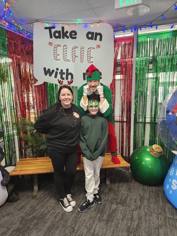 Children posing with Pinkie the Elf in front of decorations and a sign that says Take an "Elfie" with me.