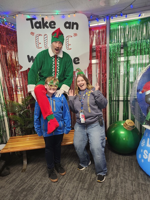 Children posing with Pinkie the Elf in front of decorations and a sign that says Take an "Elfie" with me.