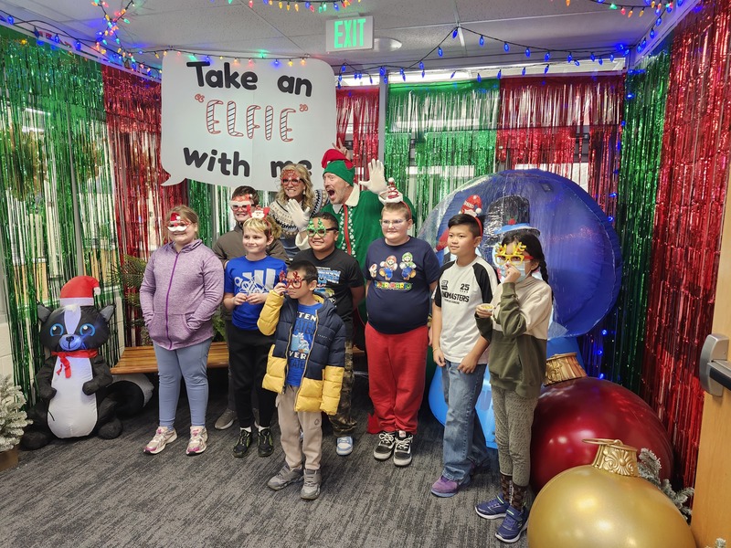 Children posing with Pinkie the Elf in front of decorations and a sign that says Take an "Elfie" with me.