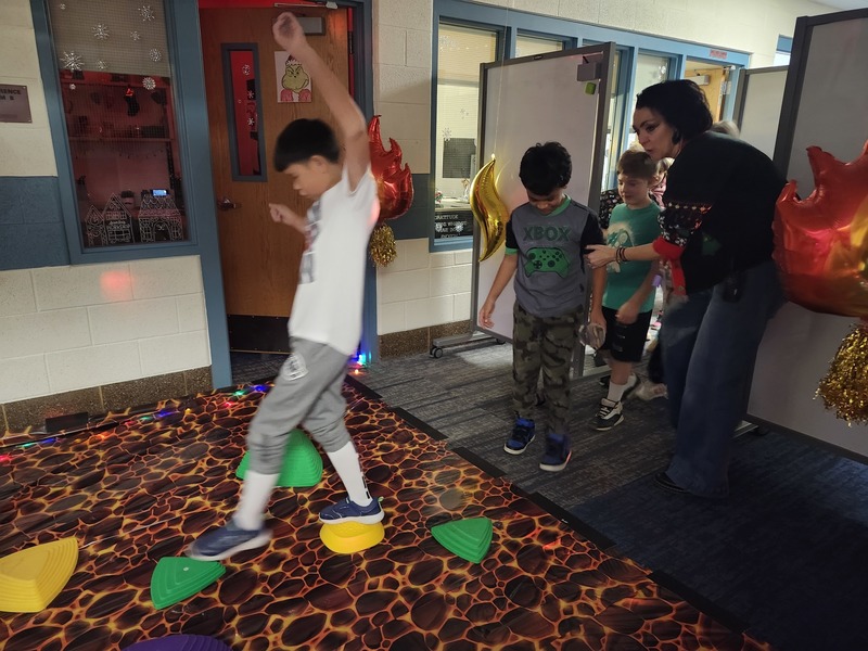 Students walking on stepping stones through a lava floor. Pinky the Elf is tied up with lights holding a sign saying help me.
