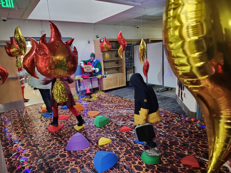 Students walking on stepping stones through a lava floor. Pinky the Elf is tied up with lights holding a sign saying help me.