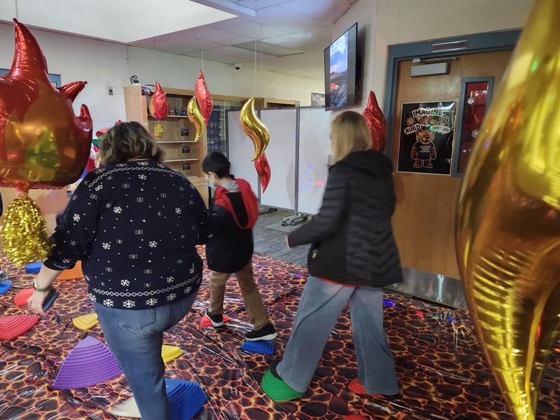 Students walking on stepping stones through a lava floor. Pinky the Elf is tied up with lights holding a sign saying help me.