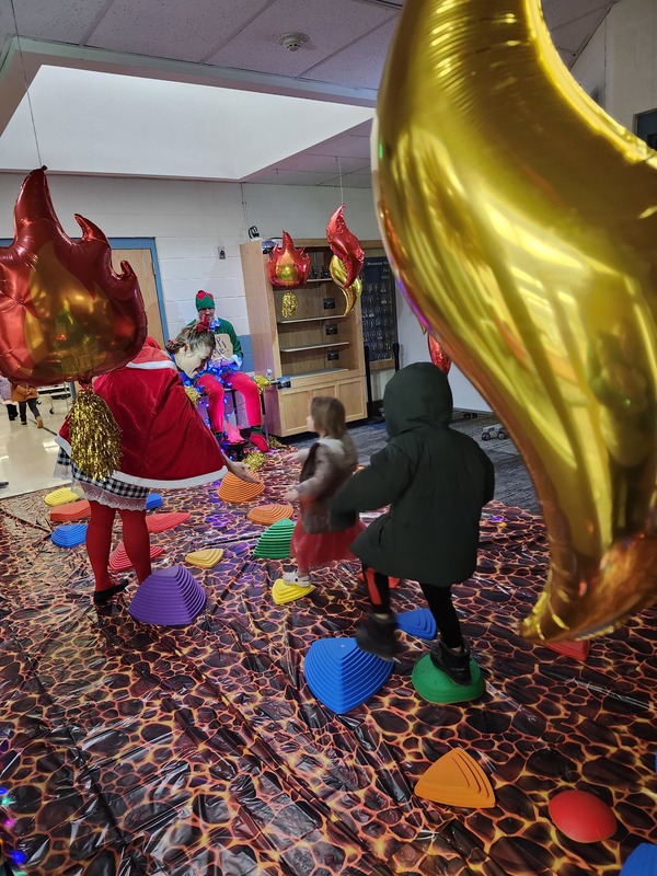 Students walking on stepping stones through a lava floor. Pinky the Elf is tied up with lights holding a sign saying help me.