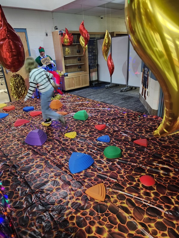 Students walking on stepping stones through a lava floor. Pinky the Elf is tied up with lights holding a sign saying help me.