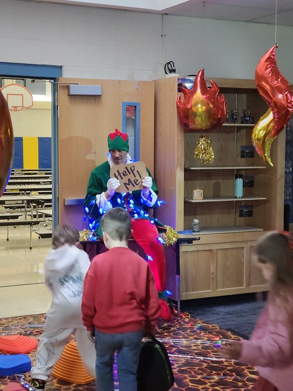 Students walking on stepping stones through a lava floor. Pinky the Elf is tied up with lights holding a sign saying help me.