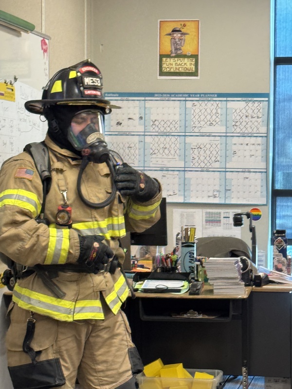 A fireman with his gear. He is showing it to students in a classroom.
