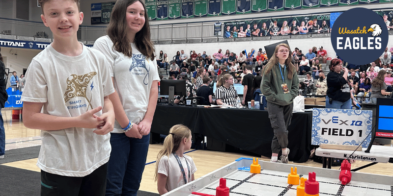 Students stand beside a VEX IQ robotics competition field inside a crowded gymnasium, with red and yellow game pieces arranged on the field, judges’ tables in the background, and a large “Wasatch Eagles” sign visible above the competition area.