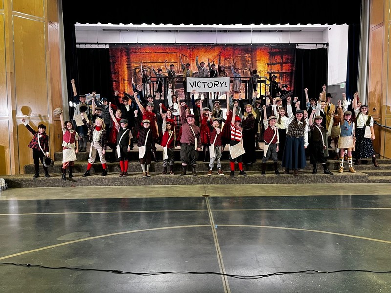 Students dressed in costume for the Newsies Jr. school play standing on the stairs in from of the play backdrop