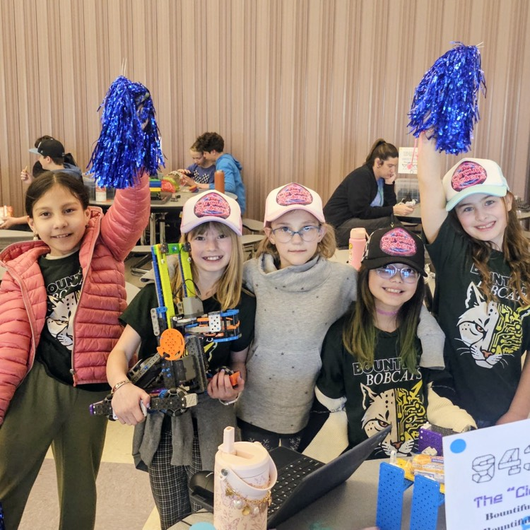 Five Bountiful Elementary students from The Circuit Sisters VEX IQ robotics team smile at their competition table, with blue pom-poms raised, a robot build on display, and team gear visible.