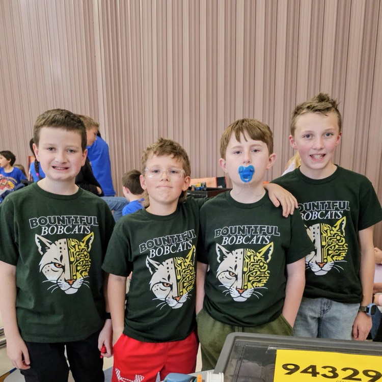 Four Bountiful Elementary students from the Baby Boomers VEX IQ robotics team stand together in matching Bobcat shirts beside their competition table at a robotics event.