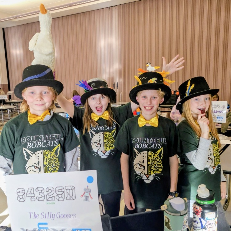 Four Bountiful elementary students from the Silly Gooses VEX IQ robotics team pose in decorated top hats and yellow bow ties beside their team sign at competition.