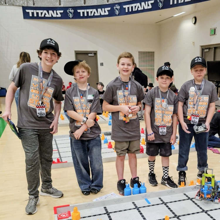 Five Bountiful Elementary students from the BOT_BROS VEX IQ robotics team stand on the competition floor in Utah State Championship shirts beside their robot on the field.
