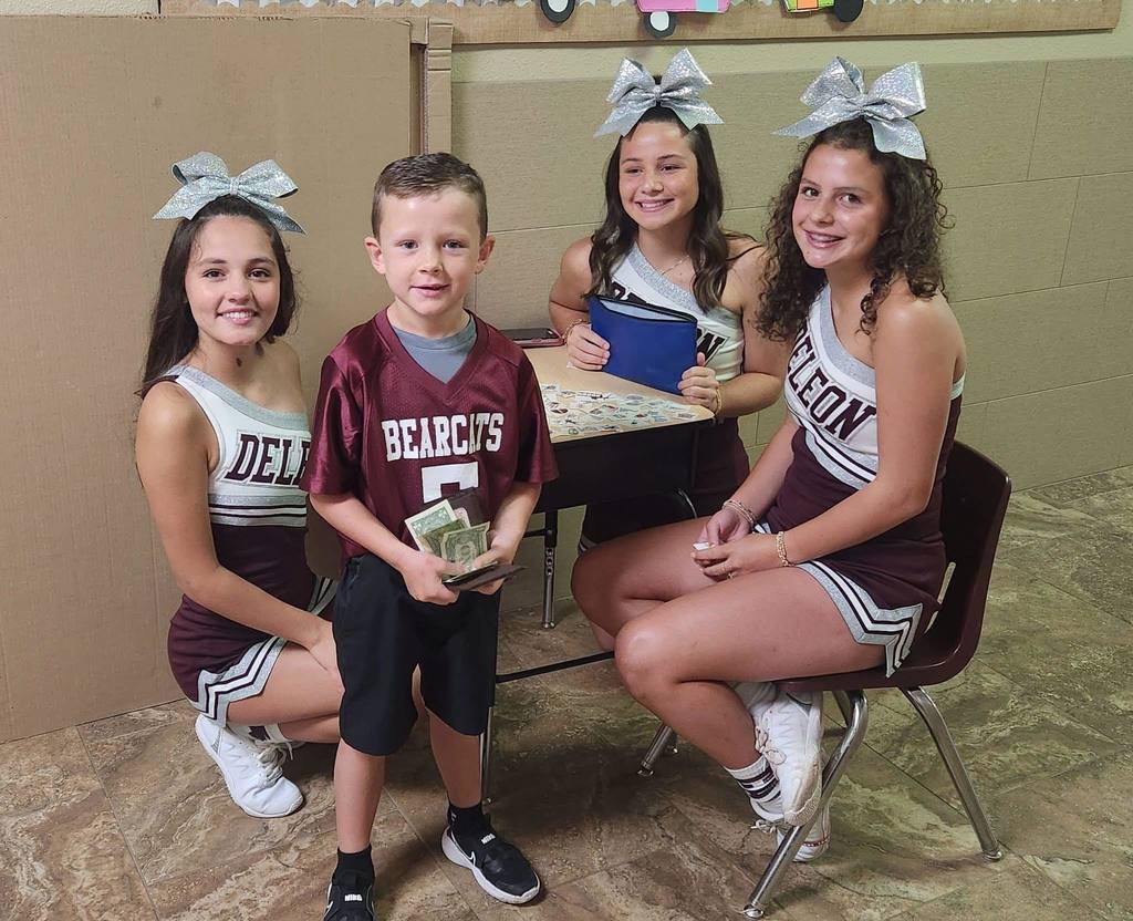 three cheerleaders sitting at desk selling tattoos to boy in bearcat jersey