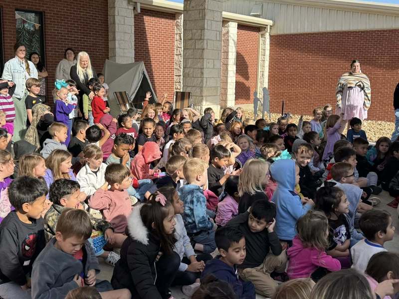 group of students sitting in front of elementary school listening to guest speaker