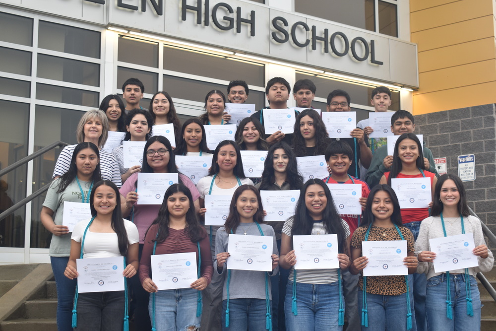 Students holding Seal of Biliteracy Certificates with turquoise graduation cords draped around their neck standing on steps in front of De Queen High School