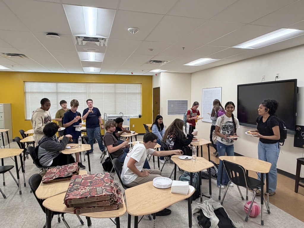 A lively classroom pizza party with diverse students standing and sitting, enjoying pizza. The room has bright lighting and a cheerful atmosphere.