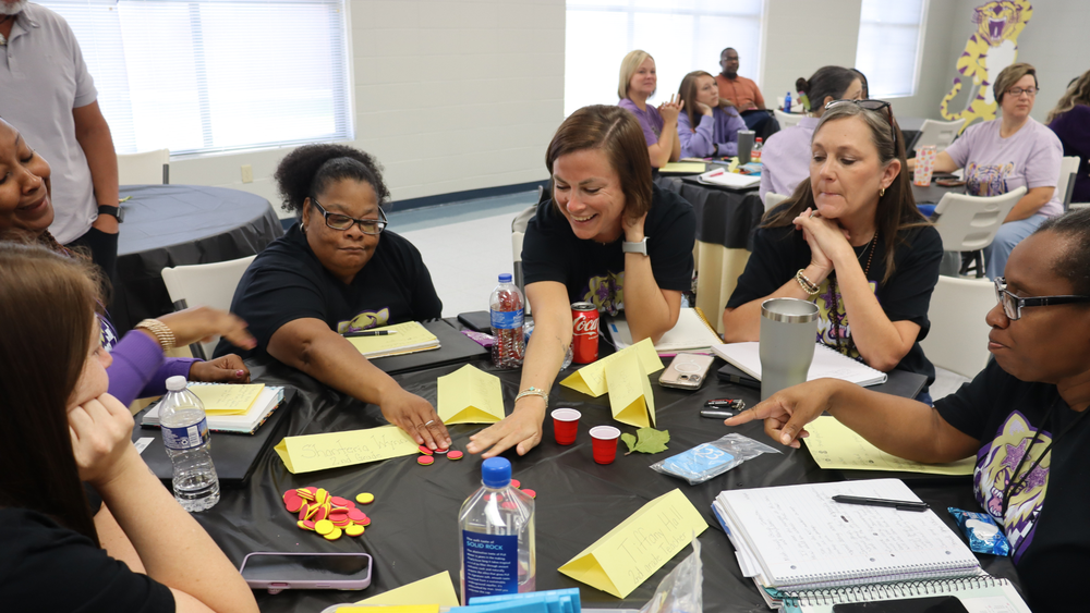 A group of educators sits around a table during a collaborative workshop, smiling and reaching toward game pieces as they participate in an interactive activity.