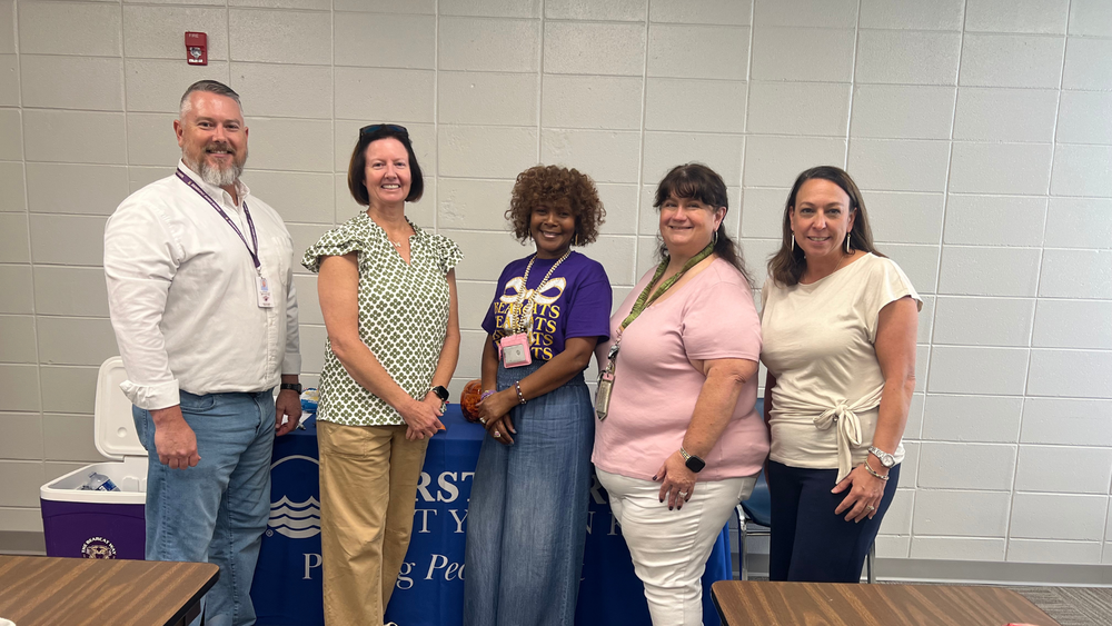 Five school staff members stand together smiling in front of a blue table decorated for an event.