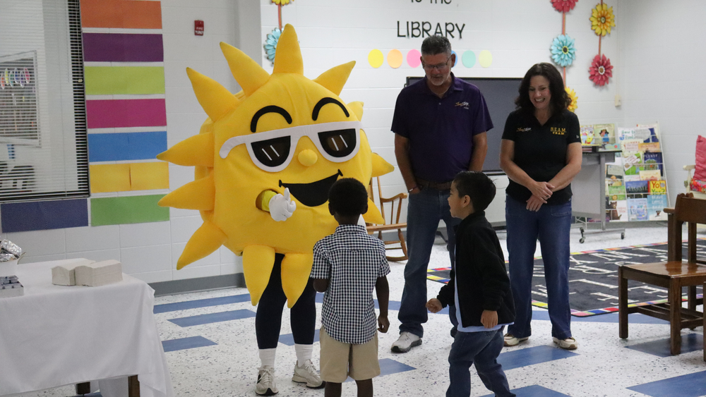 Two students greet a smiling sun mascot during a cheerful school library event.